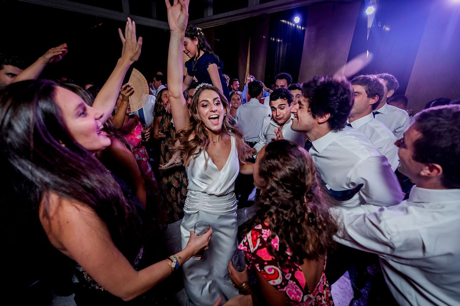 Laughing bride in white dress dancing joyfully surrounded by celebrating guests at vibrant wedding reception.