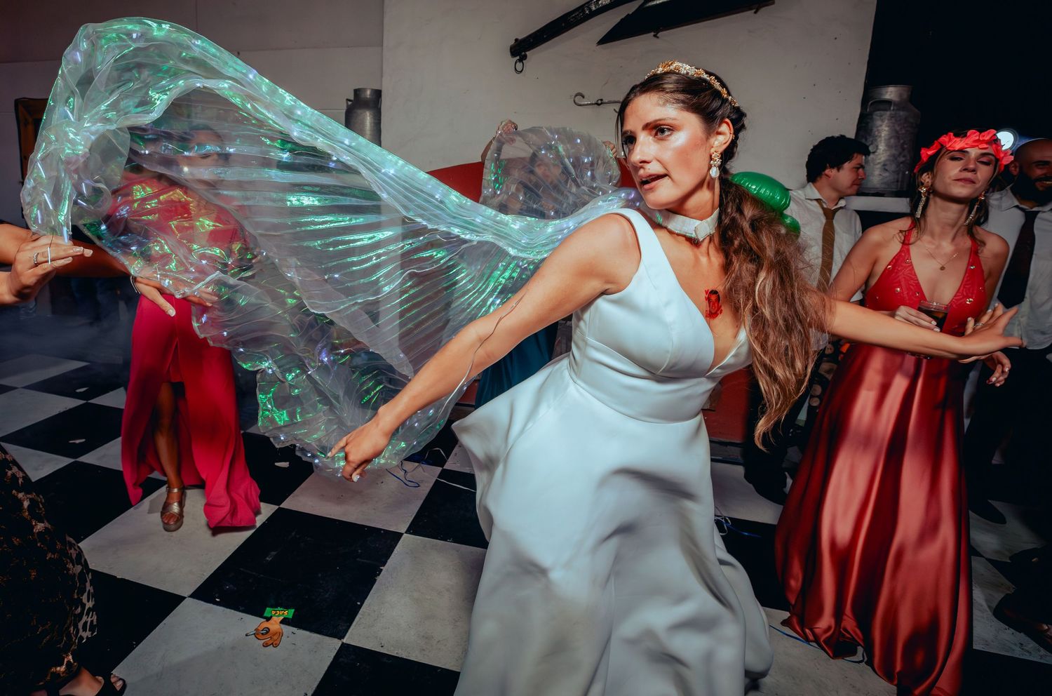 Bride twirling in white gown with iridescent wings on a checkered dance floor at wedding reception.
