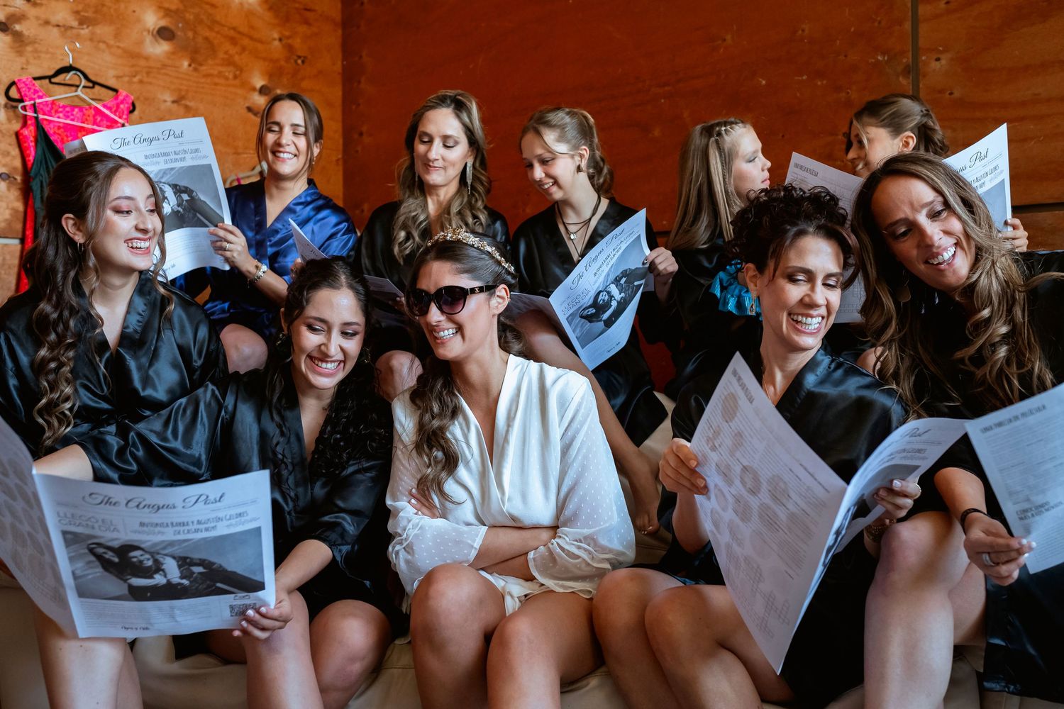 Bride in white robe with bridesmaids in black robes laughing while reading wedding newspapers together.
