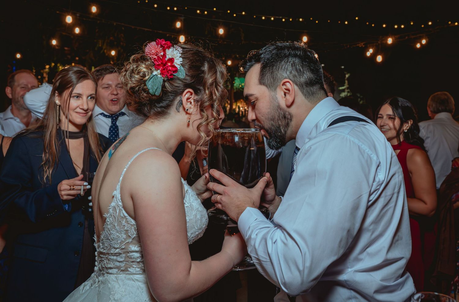 Bride and groom share intimate moment drinking from large glass at outdoor wedding.
