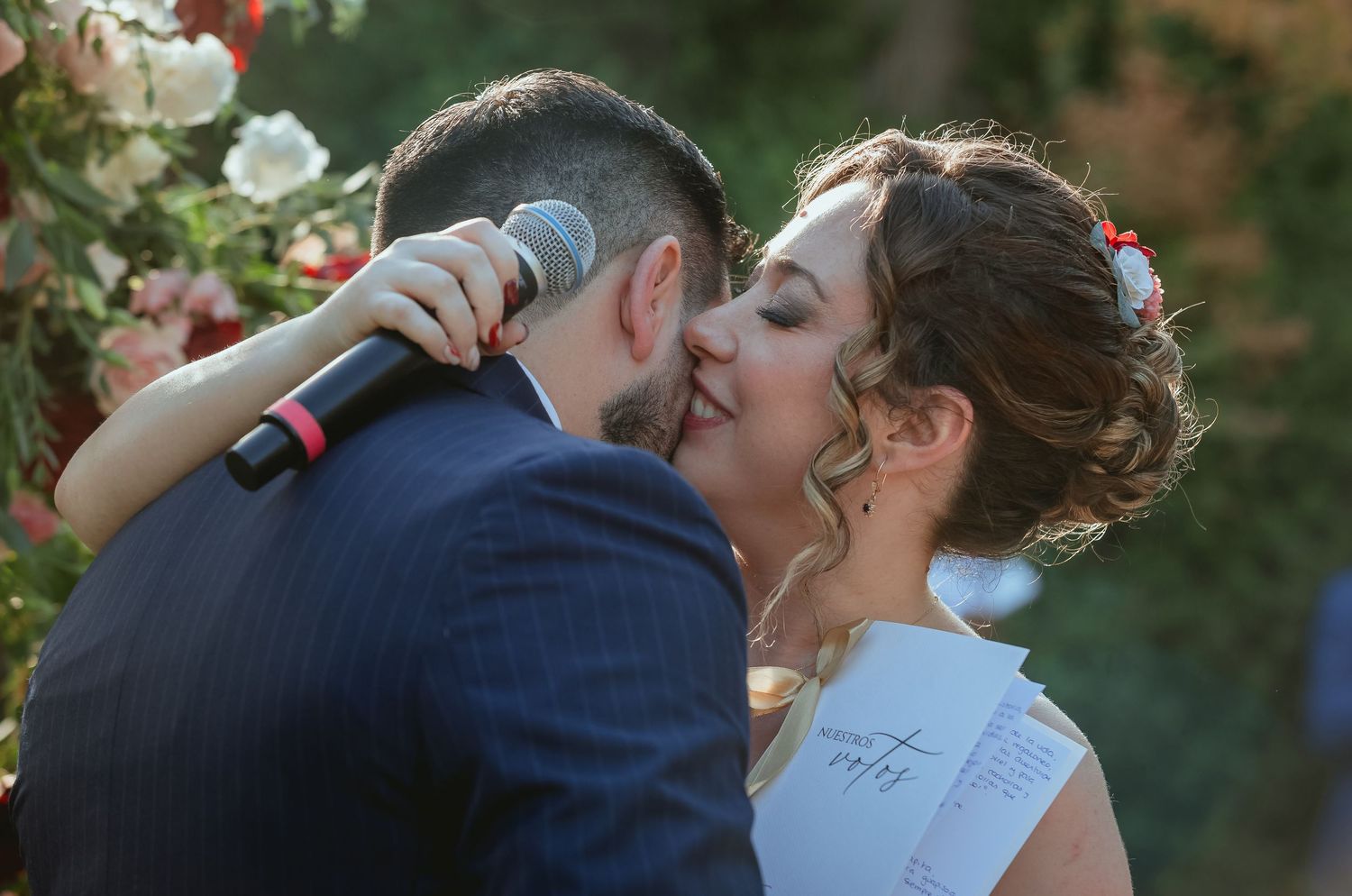 Bride and groom share their first kiss at outdoor wedding ceremony while groom holds microphone.