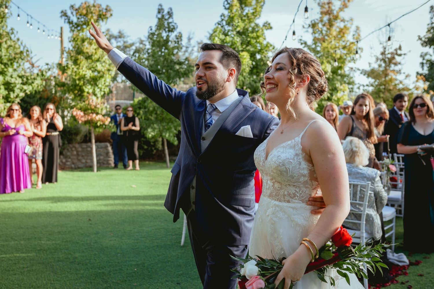 Smiling groom waves while bride holds bouquet as they exit outdoor wedding ceremony on green lawn.