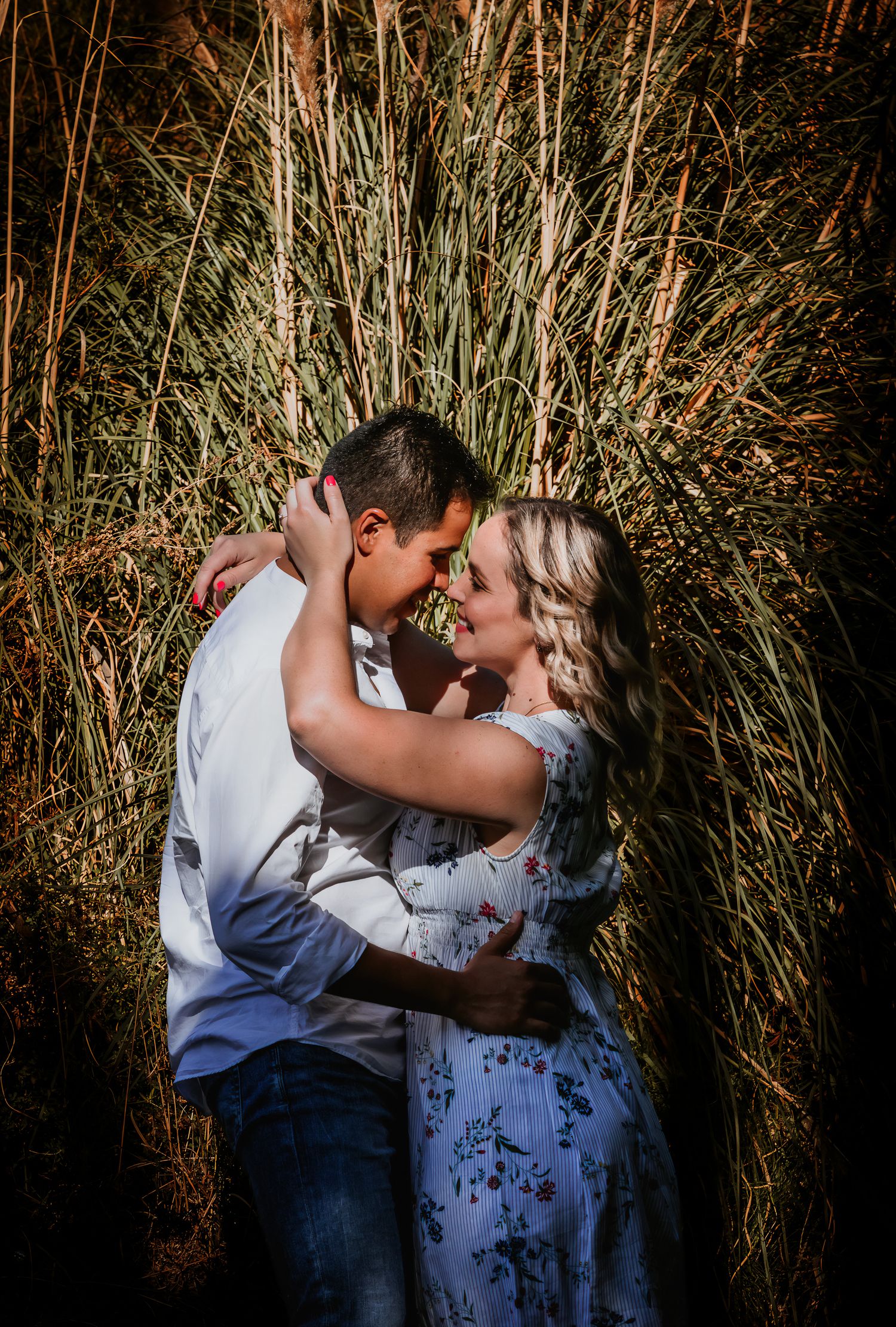 Couple embracing and almost kissing in front of tall green grass during an outdoor engagement photo session.