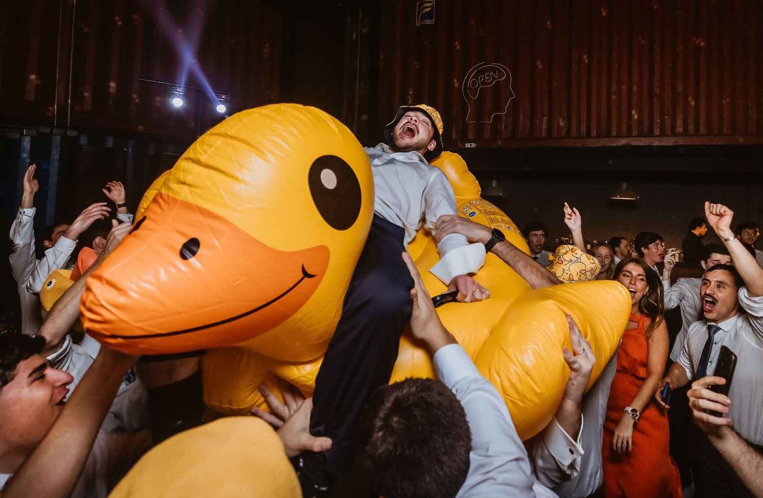 Groom crowd surfing on giant inflatable yellow rubber duck at lively wedding reception party.