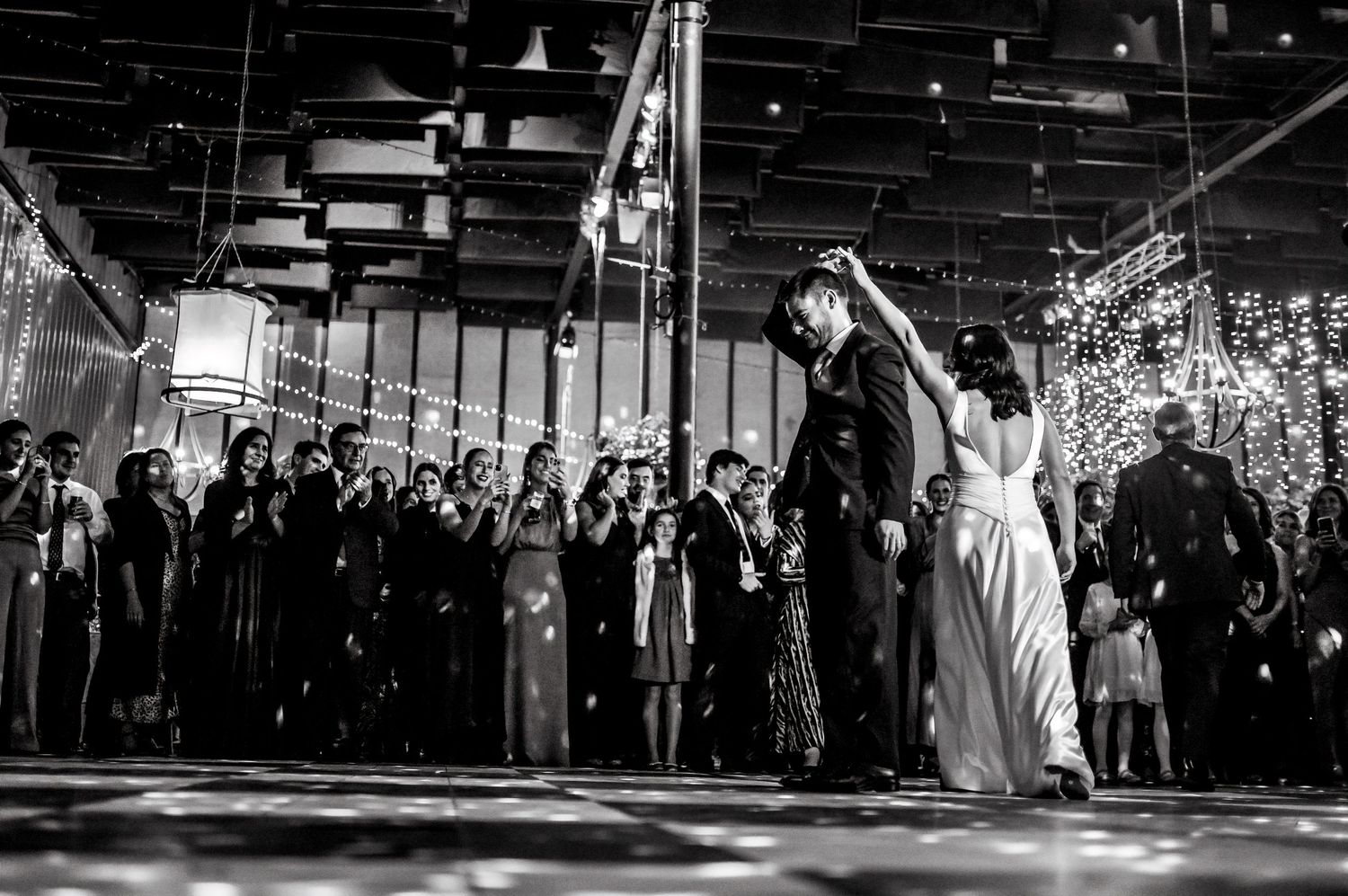 Black and white photo of a couple sharing their first dance at a wedding reception surrounded by guests and string lights.