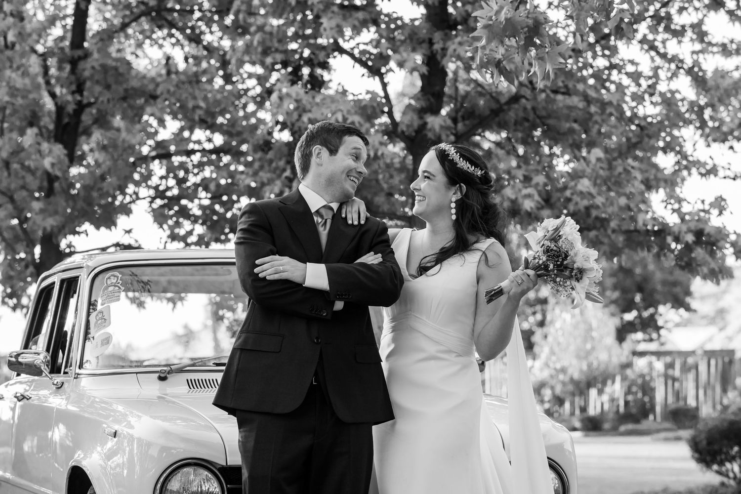 Black and white wedding photo of smiling bride and groom standing beside a vintage car outdoors.