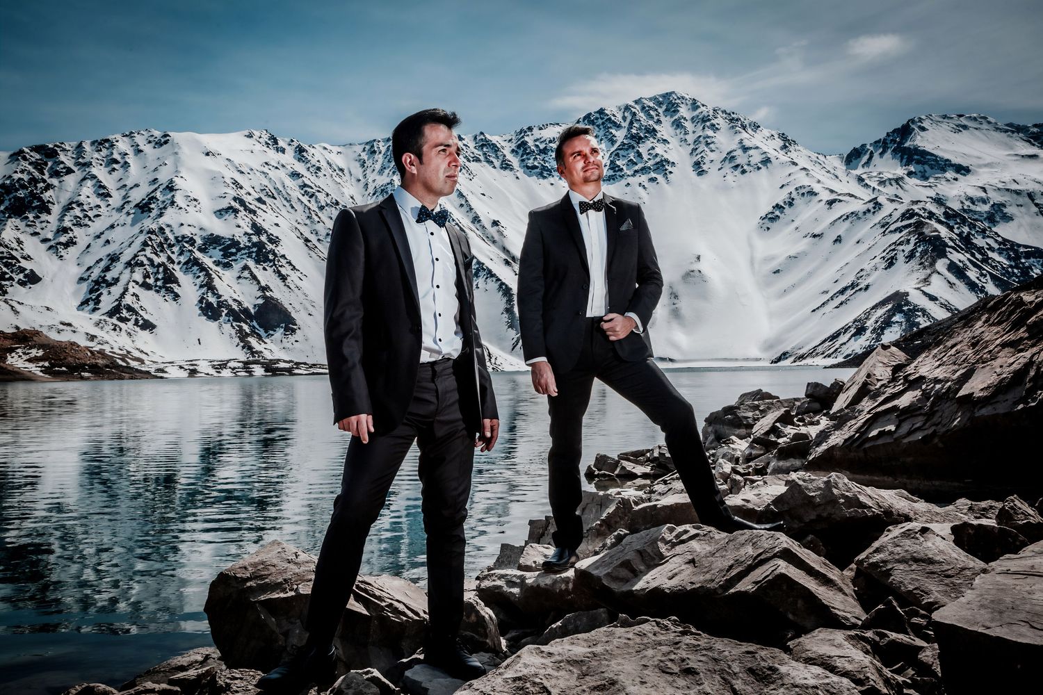 Two men in tuxedos pose on rocks before a stunning snow-capped mountain lake backdrop.