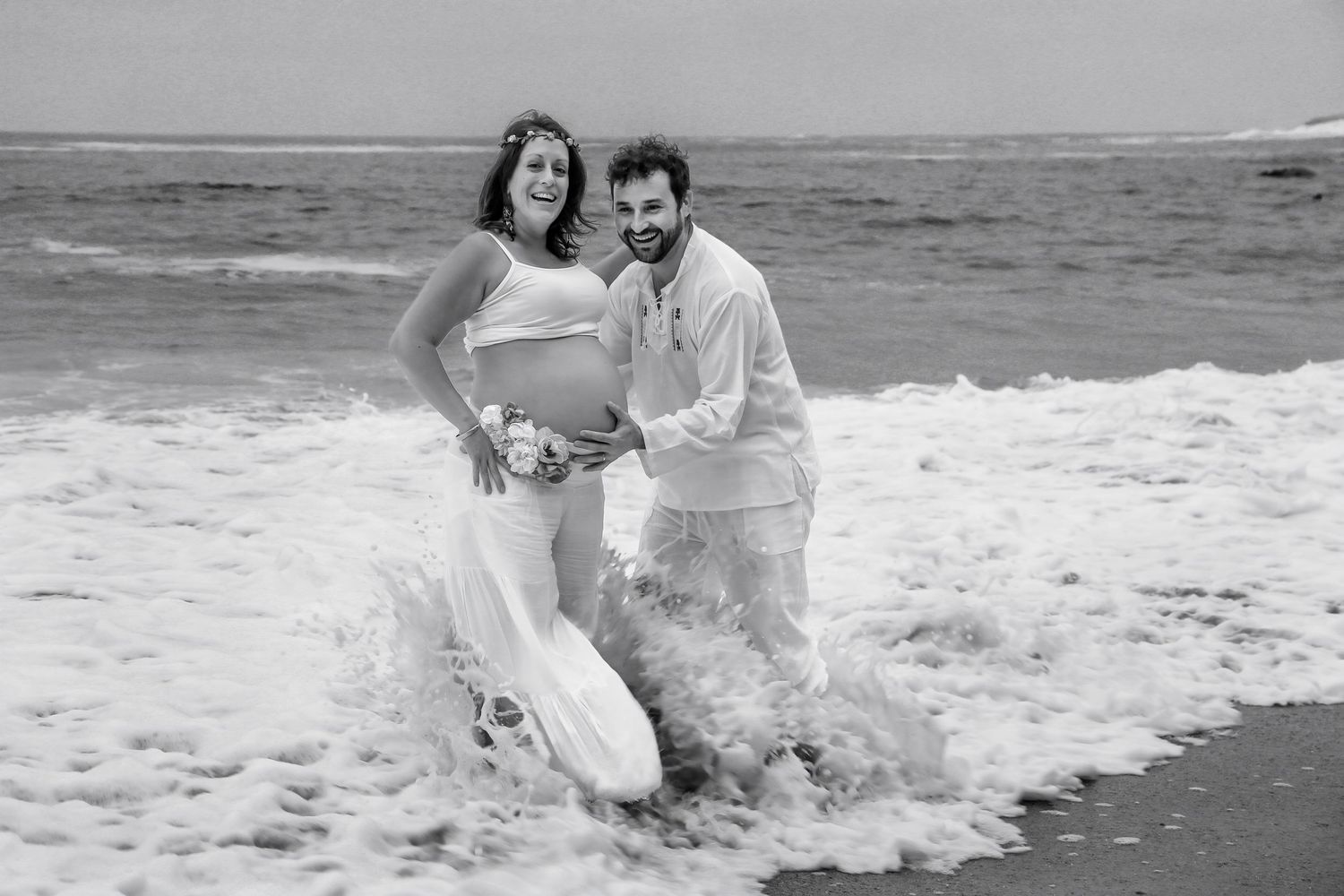 Black and white photo of a pregnant couple laughing as ocean waves splash around them on a beach.