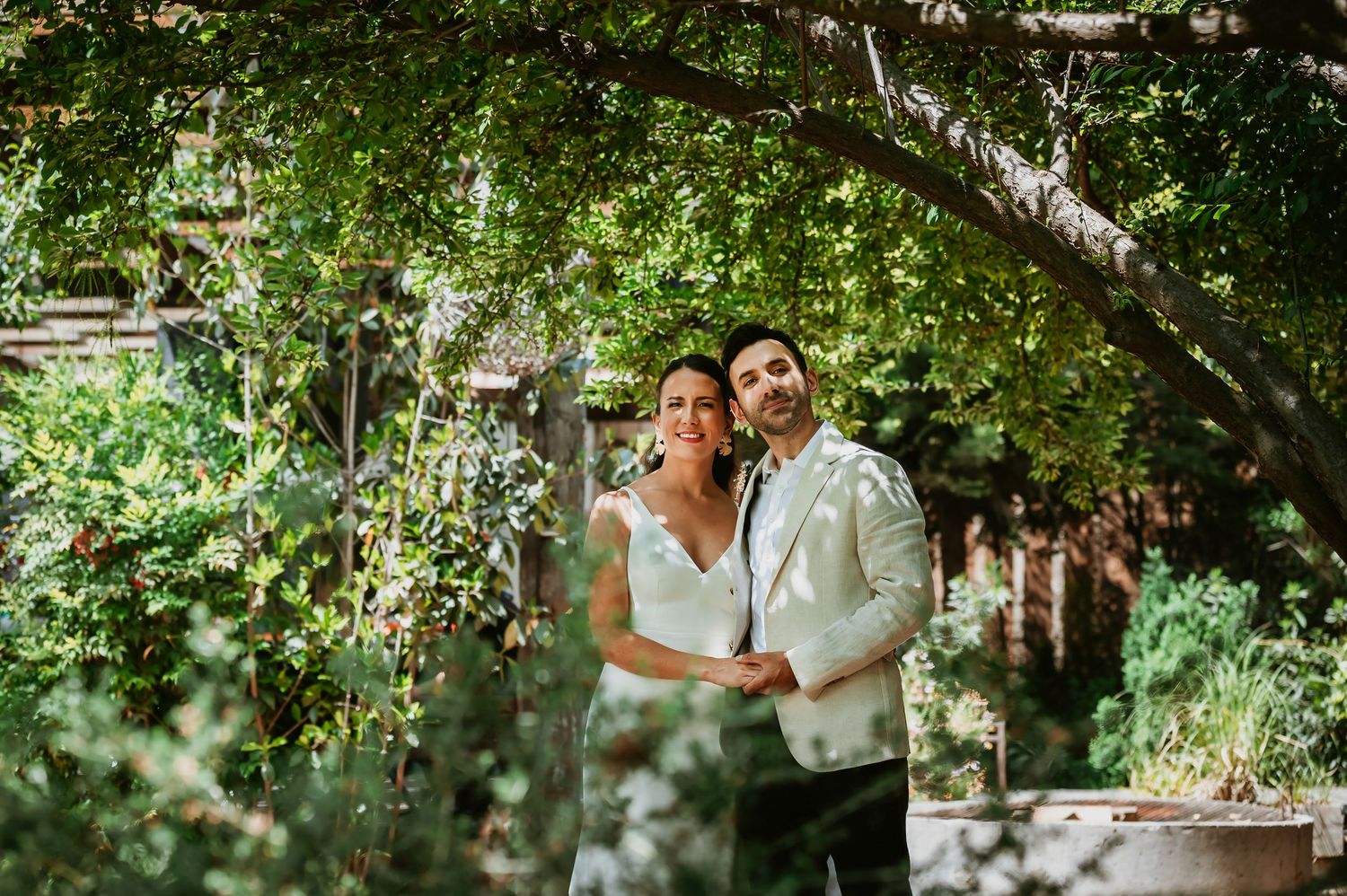 A couple embraces under a lush green tree canopy in a garden, dressed in wedding attire.