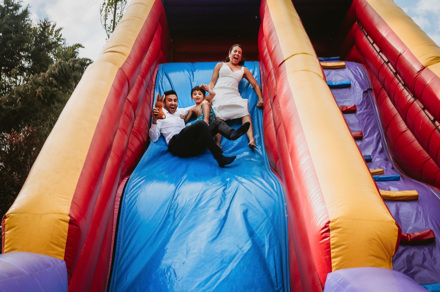 Three people laughing and sliding down a colorful inflatable bounce house slide outdoors.