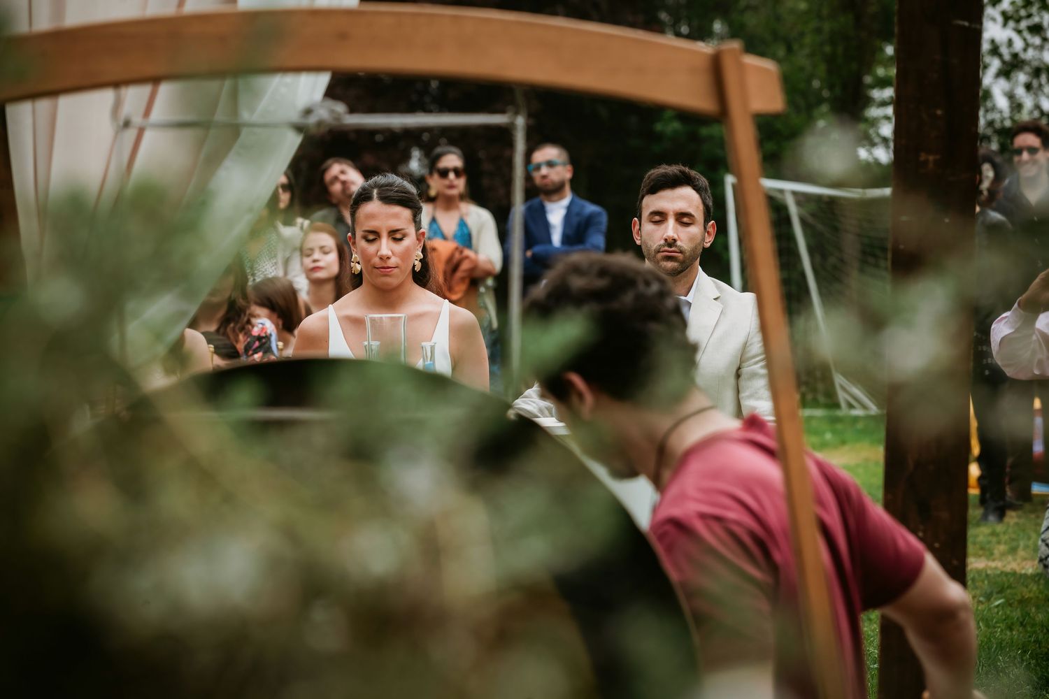 Outdoor wedding ceremony with bride in white dress and guests gathered under wooden arch surrounded by greenery.