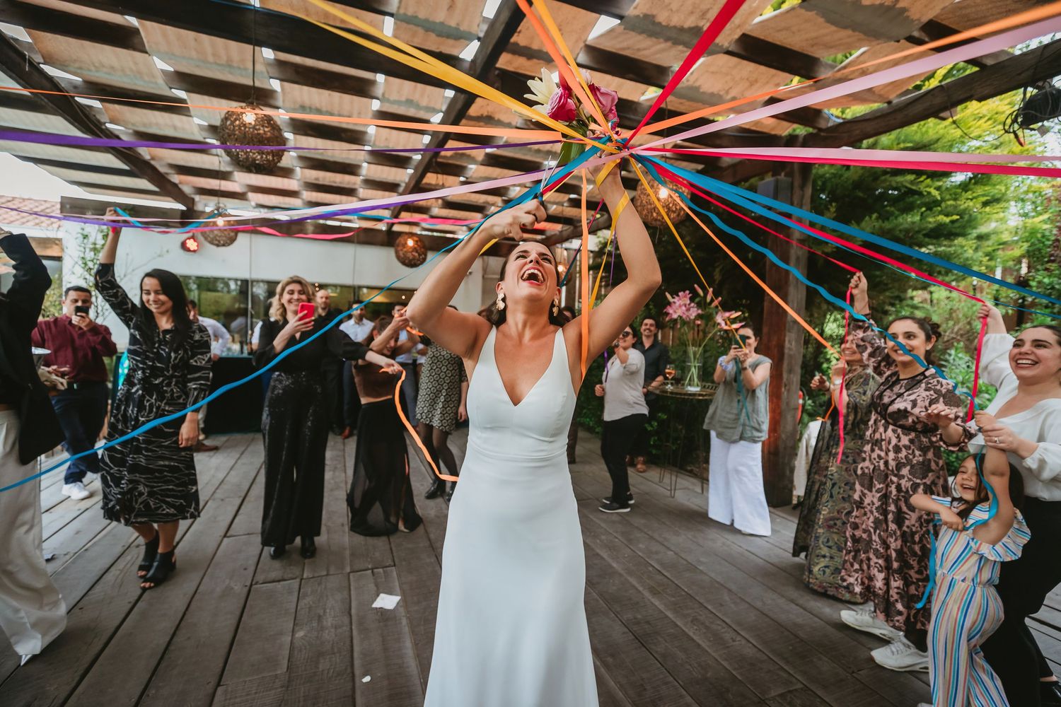 Bride in white dress joyfully tosses colorful ribbons at outdoor wedding reception with guests celebrating around her.