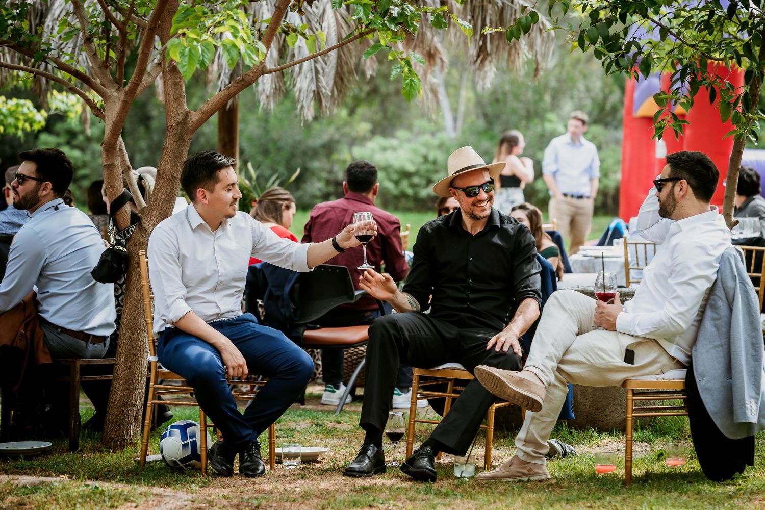 Men socializing outdoors at a garden party, raising glasses in a toast among lush green trees.