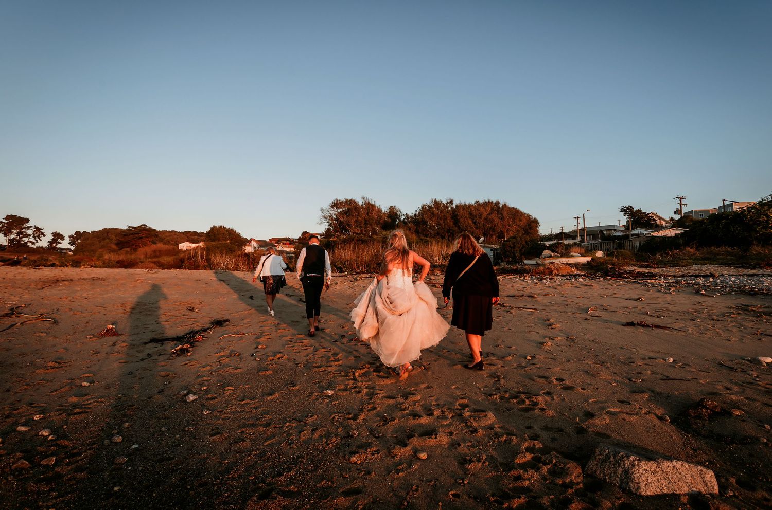 Wedding party walking on sandy beach at golden hour sunset with warm light casting long shadows.