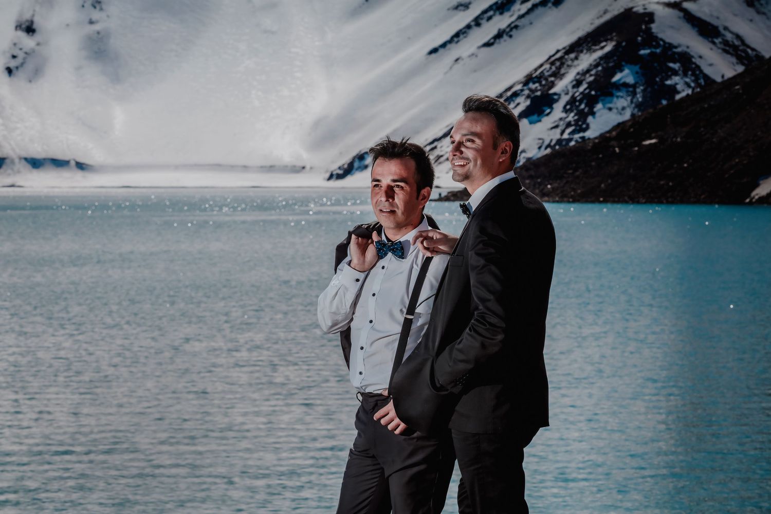 Two grooms in formal wear smile together with a glacial blue lake and snowy mountains behind.