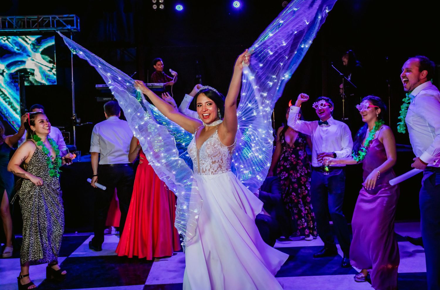 Bride in white gown dancing with glowing LED butterfly wings on a crowded wedding dance floor.