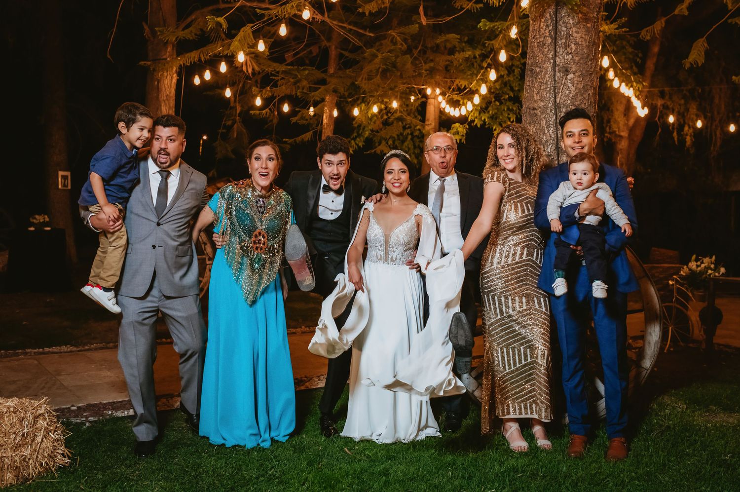 Wedding couple poses with family members outdoors at night under romantic string lights in trees.
