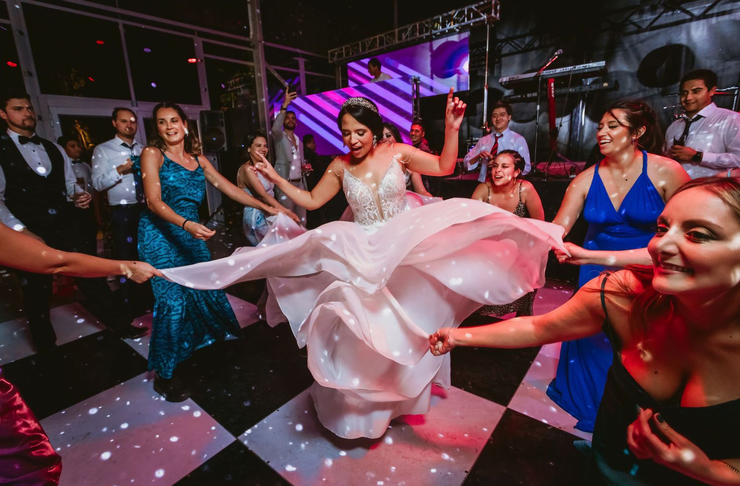 Bride in white gown twirls joyfully on checkered dance floor surrounded by guests at wedding reception.