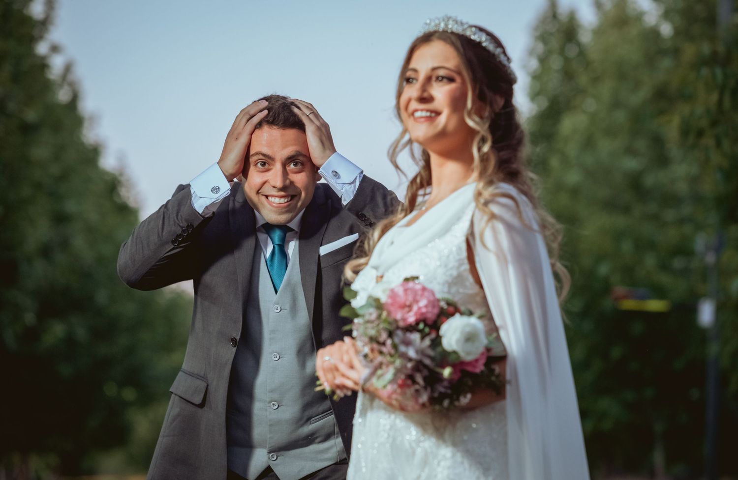 Groom makes a surprised expression beside his smiling bride holding a pink and white bouquet.