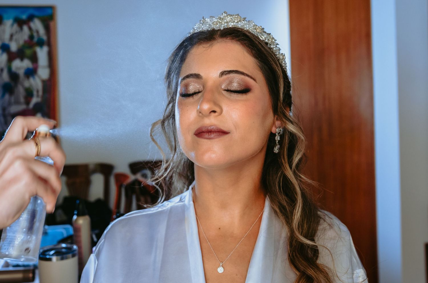 Makeup artist setting a brides face with spray while she wears a tiara and white robe.