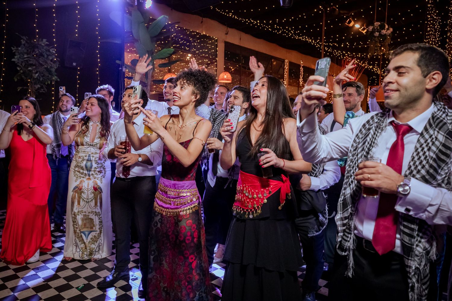Wedding guests holding phones and drinks, dancing and celebrating at a festive reception party.