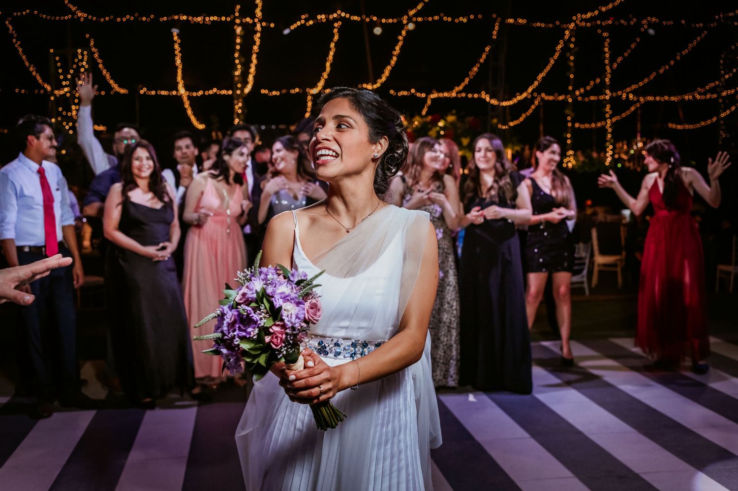 Smiling bride in white dress holds purple bouquet at an elegant wedding reception.