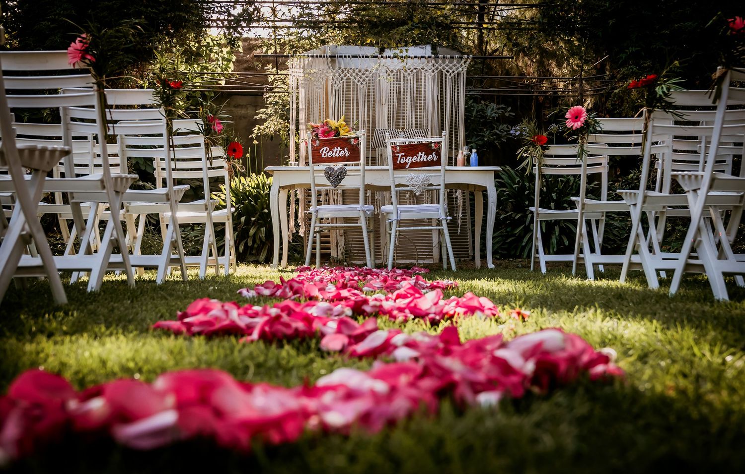 Outdoor wedding ceremony setup with white chairs, pink rose petal aisle, and macrame backdrop with Together signs.