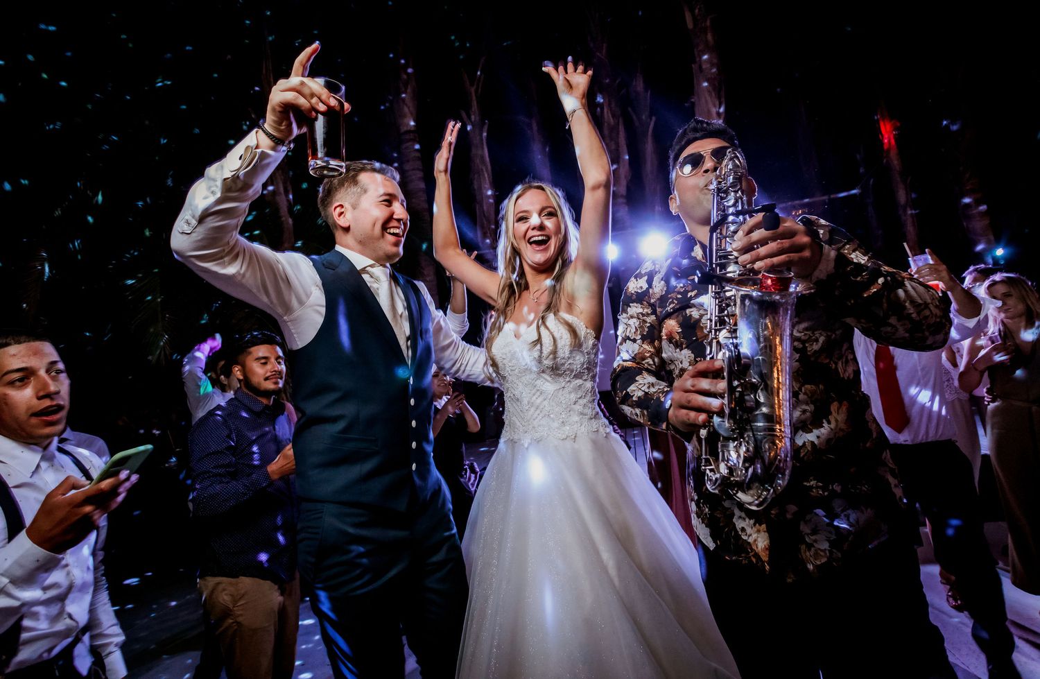 Bride and groom celebrate joyfully on wedding dance floor with saxophonist and confetti.