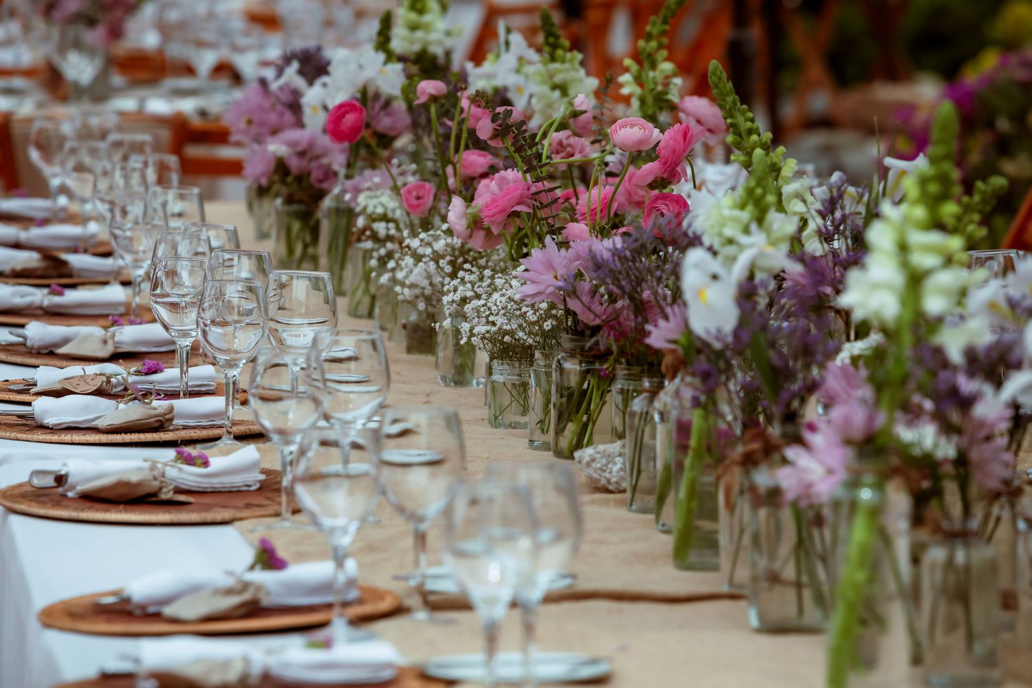 Long outdoor reception table adorned with pink and purple floral arrangements in glass vases.