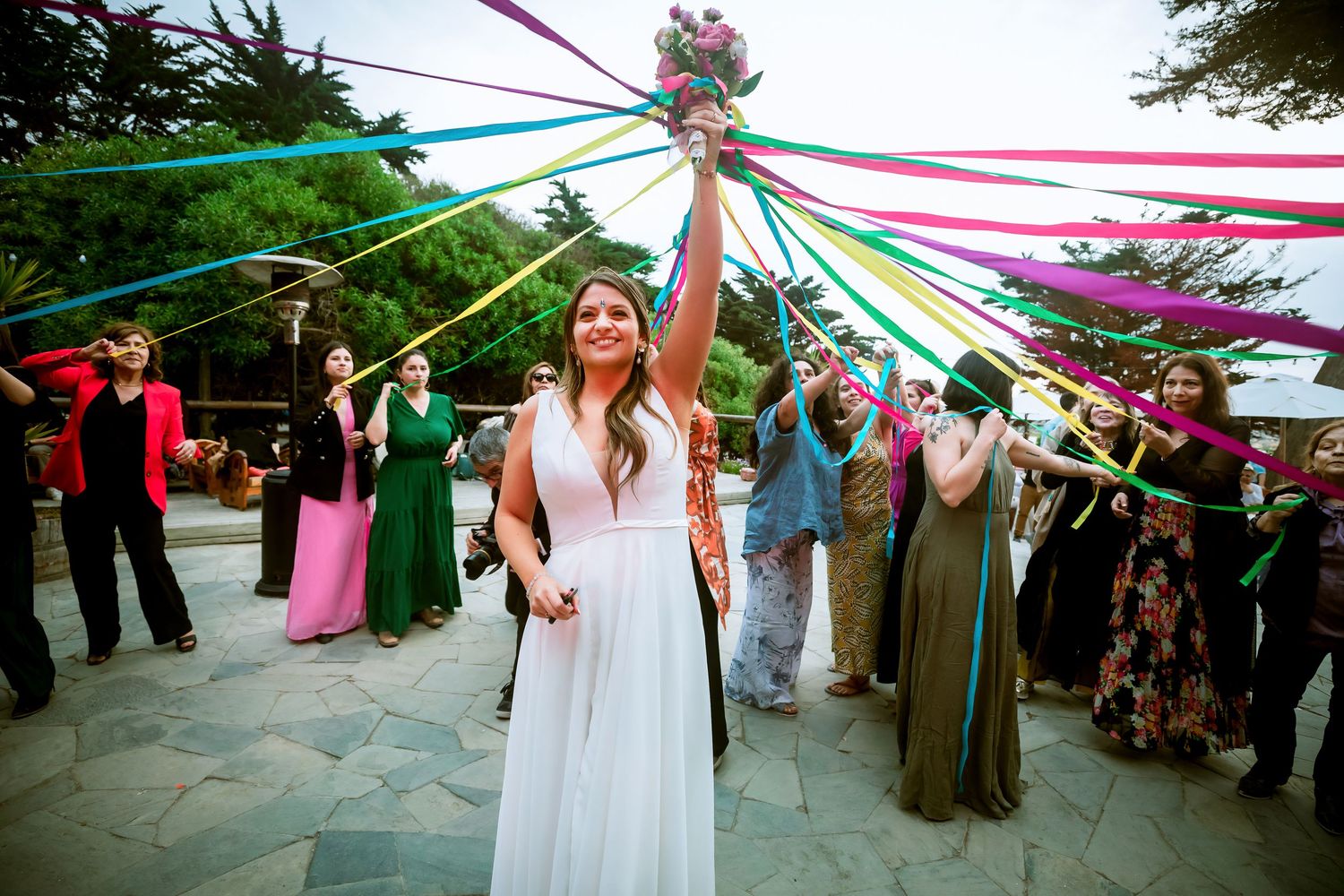 Bride tosses colorful ribbon bouquet surrounded by guests at outdoor wedding celebration.