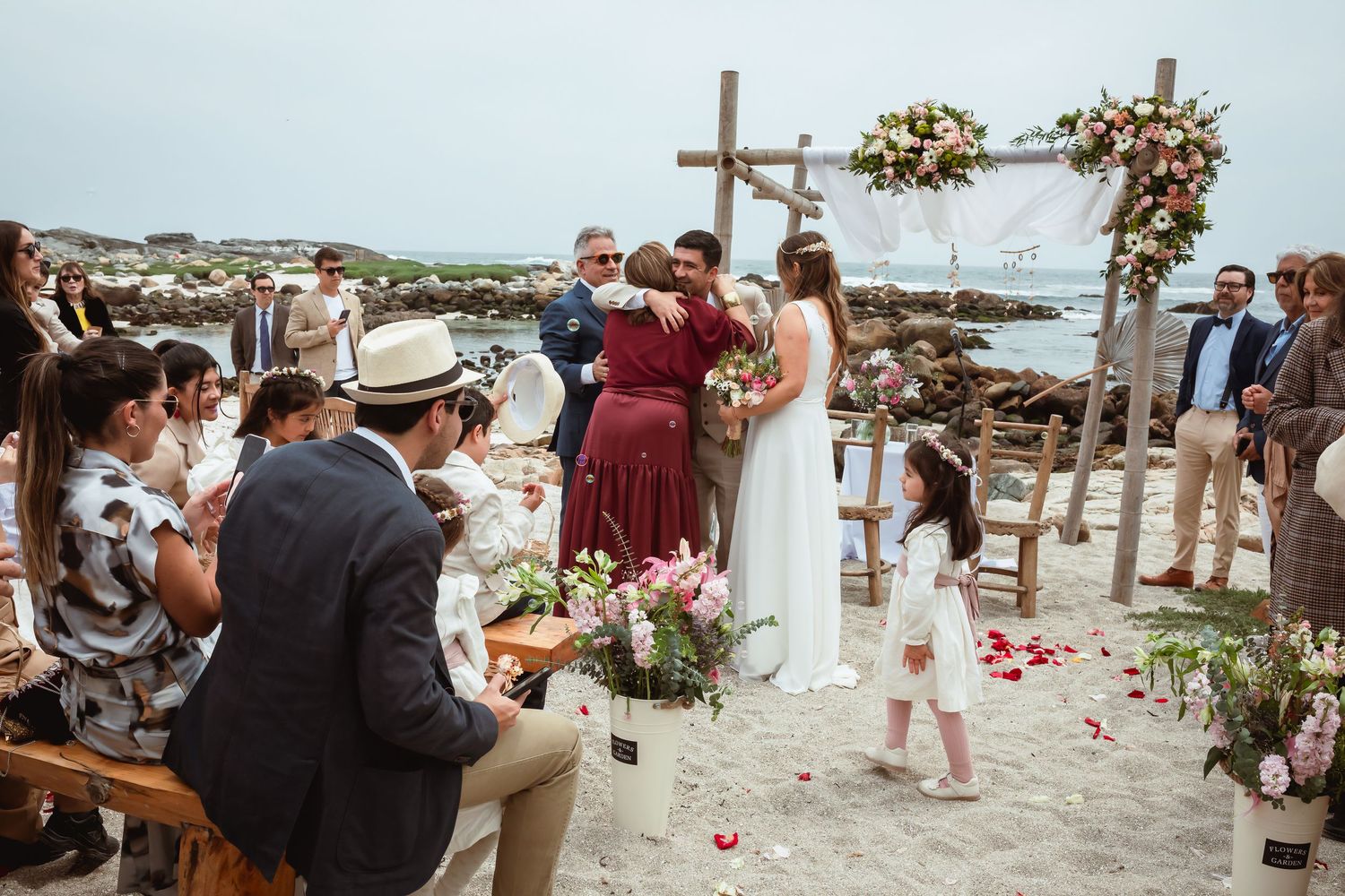 Wedding ceremony on a sandy beach with floral arch, guests seated, and a young flower girl nearby.