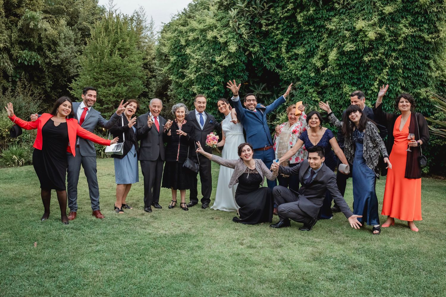 Large wedding party group photo with guests waving and celebrating on lush green lawn.