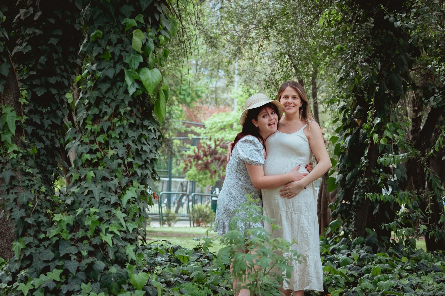 Two smiling women pose together in a green ivy-covered garden, one pregnant in a white dress.