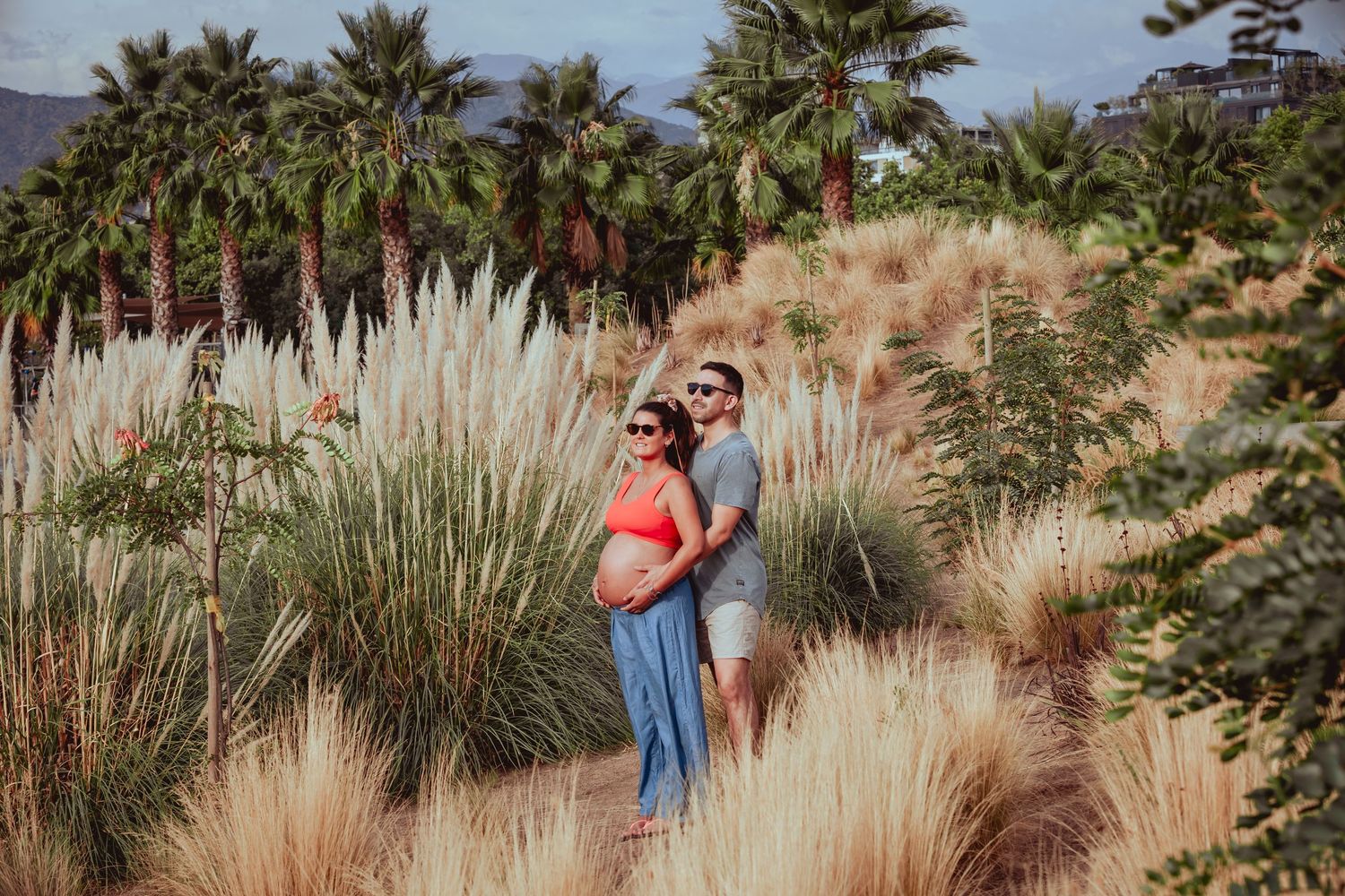 Pregnant couple embracing among tall pampas grass and palm trees during outdoor maternity photoshoot.