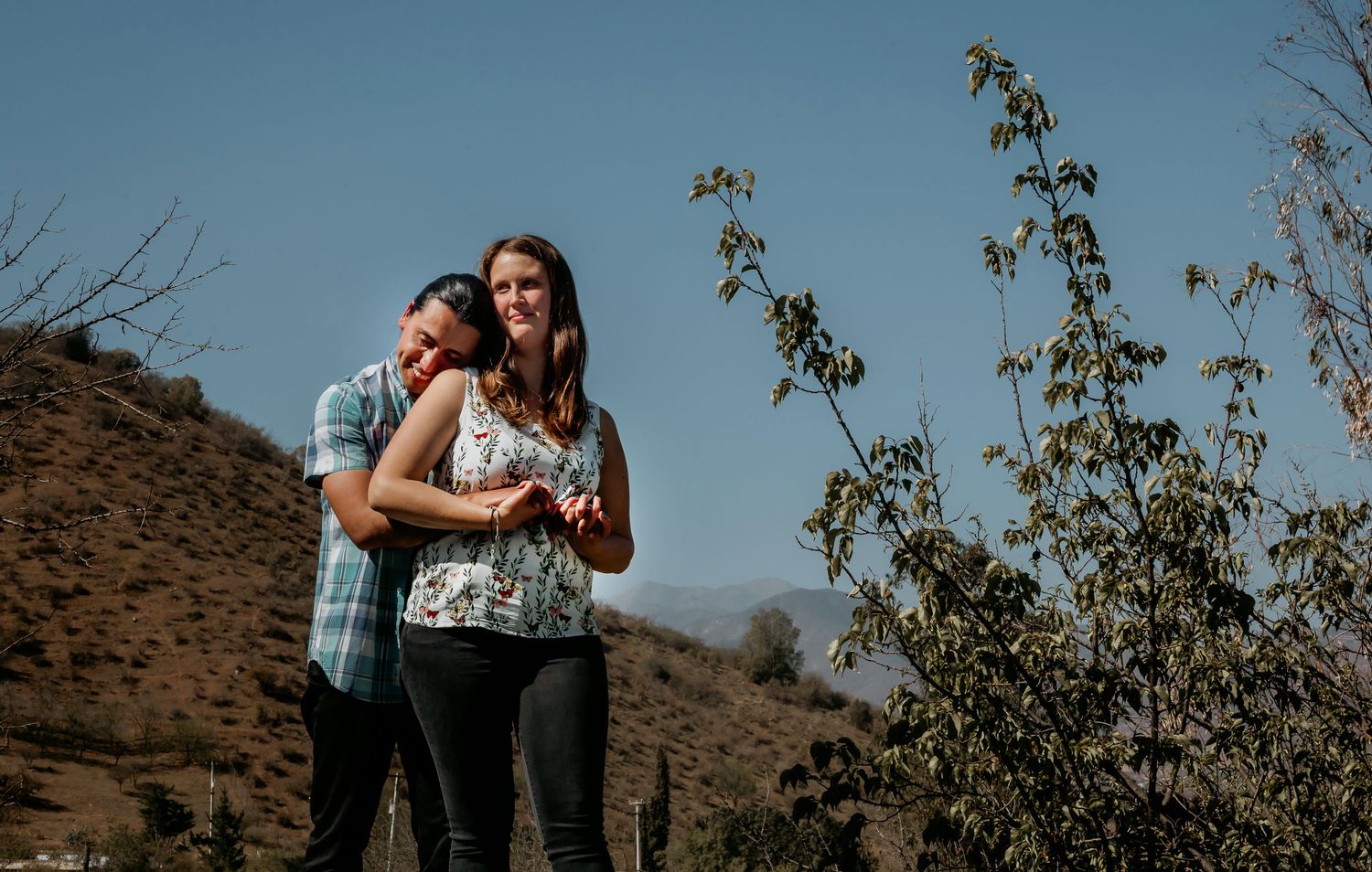 Joyful couple embracing outdoors on a sunny hillside with dry brush and mountains in the background.