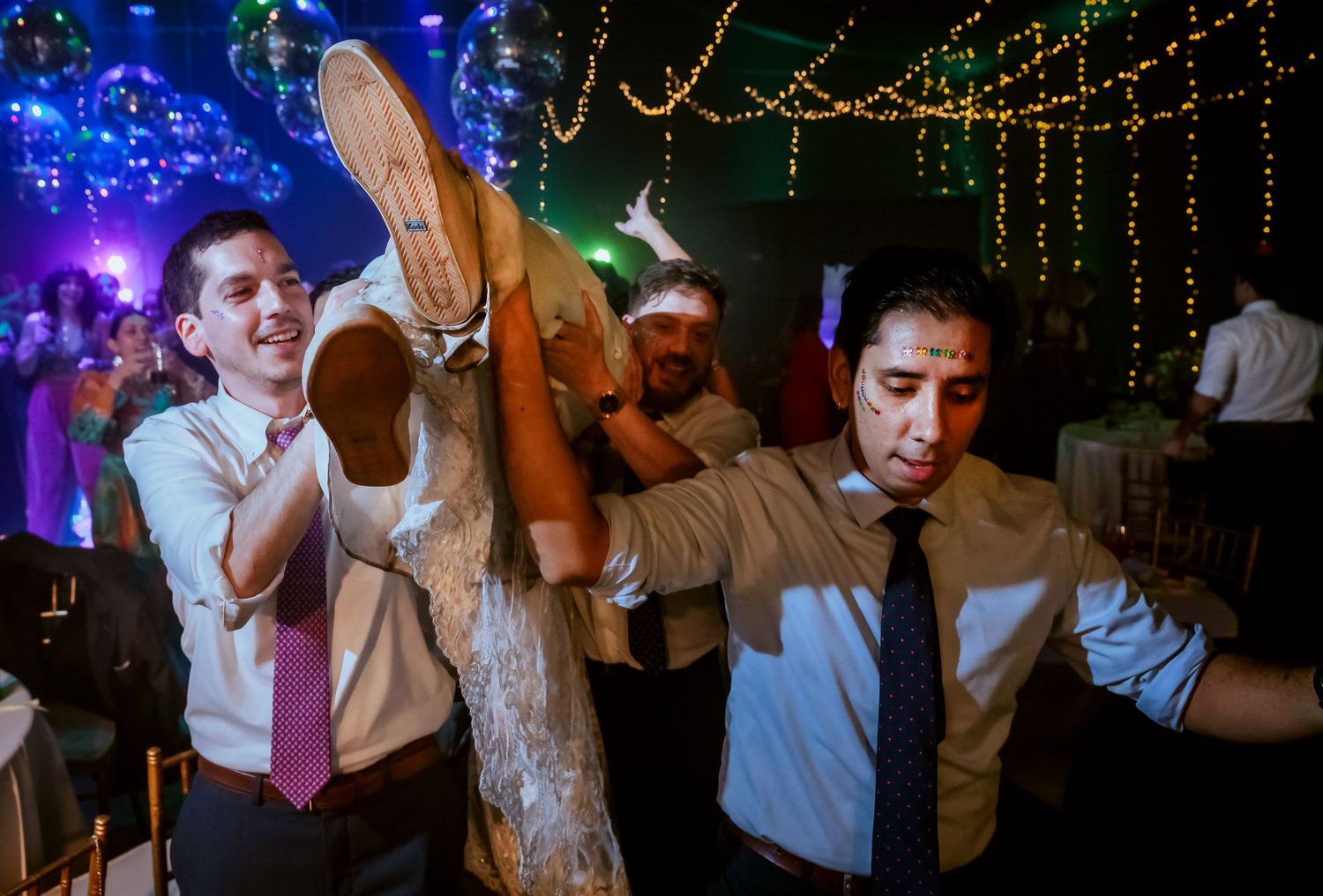 Men in dress shirts celebrate at a festive party, one lifting a tree branch while dancing under twinkling lights.