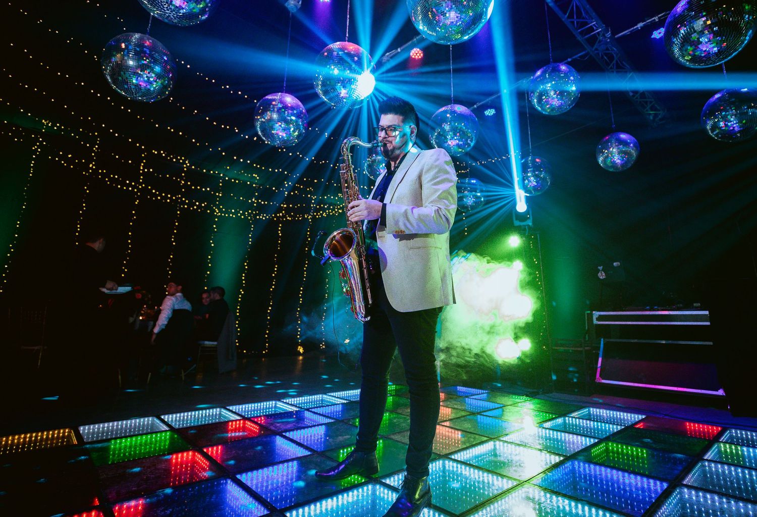 Saxophonist in white blazer performing on colorful LED dance floor under disco balls in vibrant nightclub.