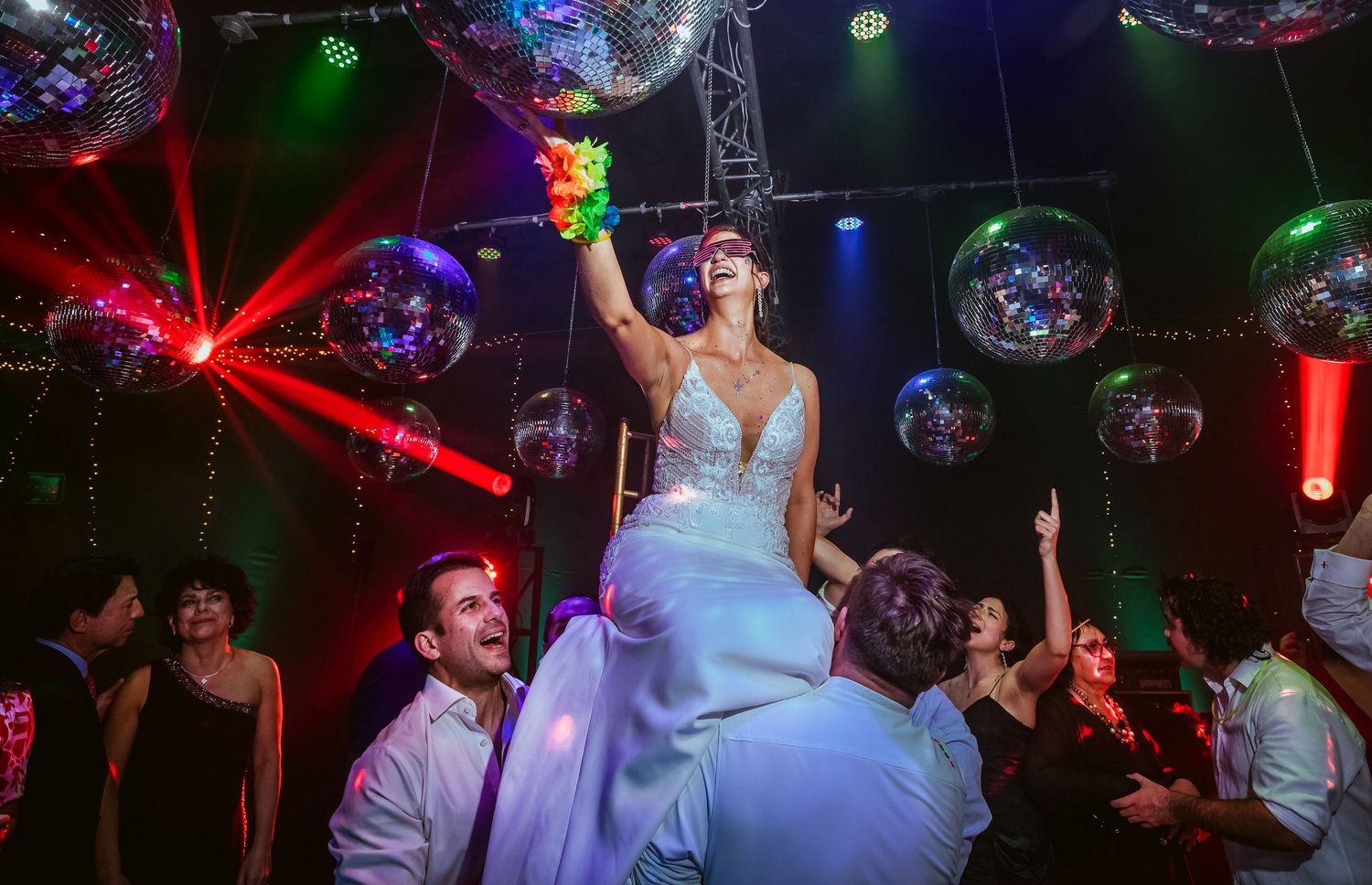 Bride in white gown lifted by guests on a vibrant dance floor with disco balls and colorful lights at a wedding reception.