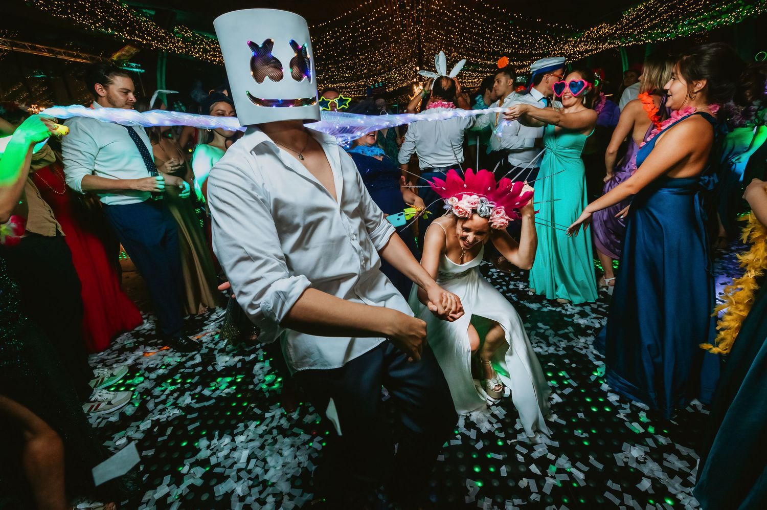Groom in Marshmello mask dips bride in white dress on confetti-covered dance floor at wedding reception.