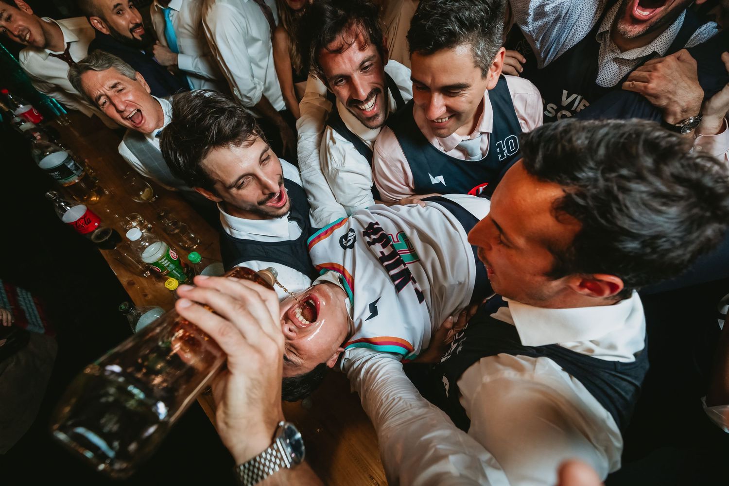Groomsmen laughing and lifting guest in cycling jersey at fun indoor wedding reception bar.