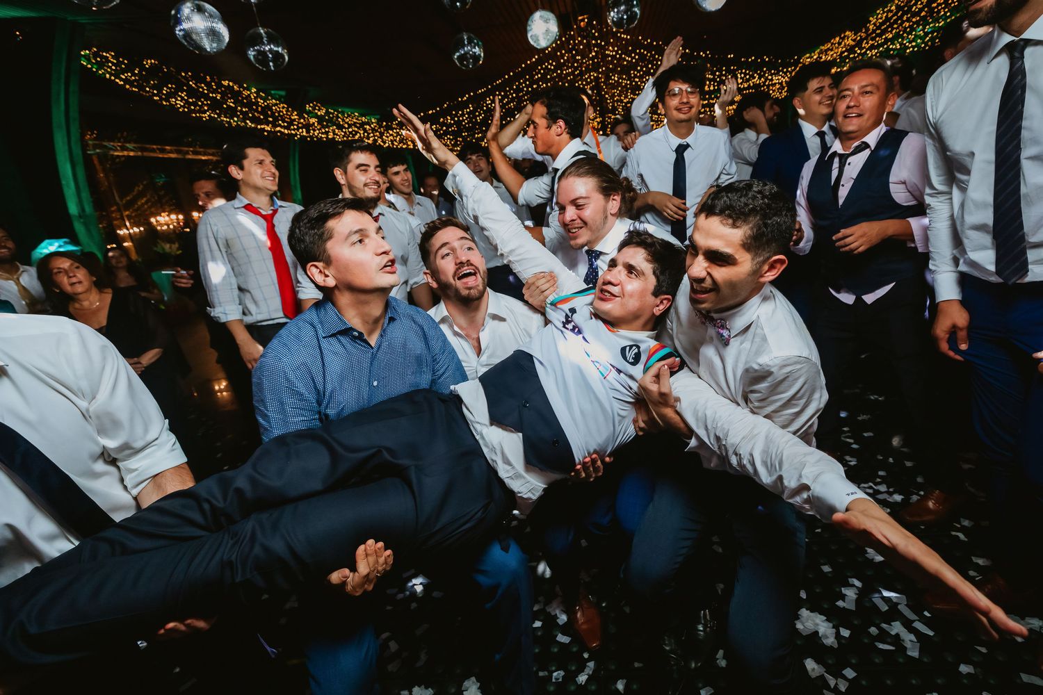 Groom lifted by cheering guests on confetti-filled wedding reception dance floor.