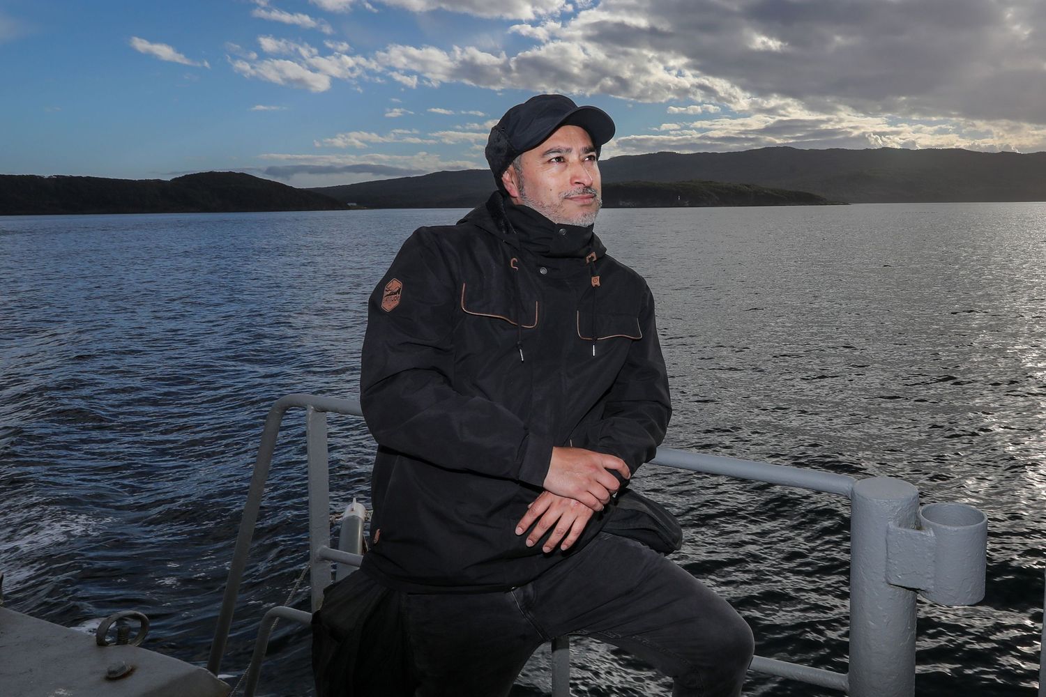 Man in black cap and jacket sitting on boat with calm lake and hills behind him.
