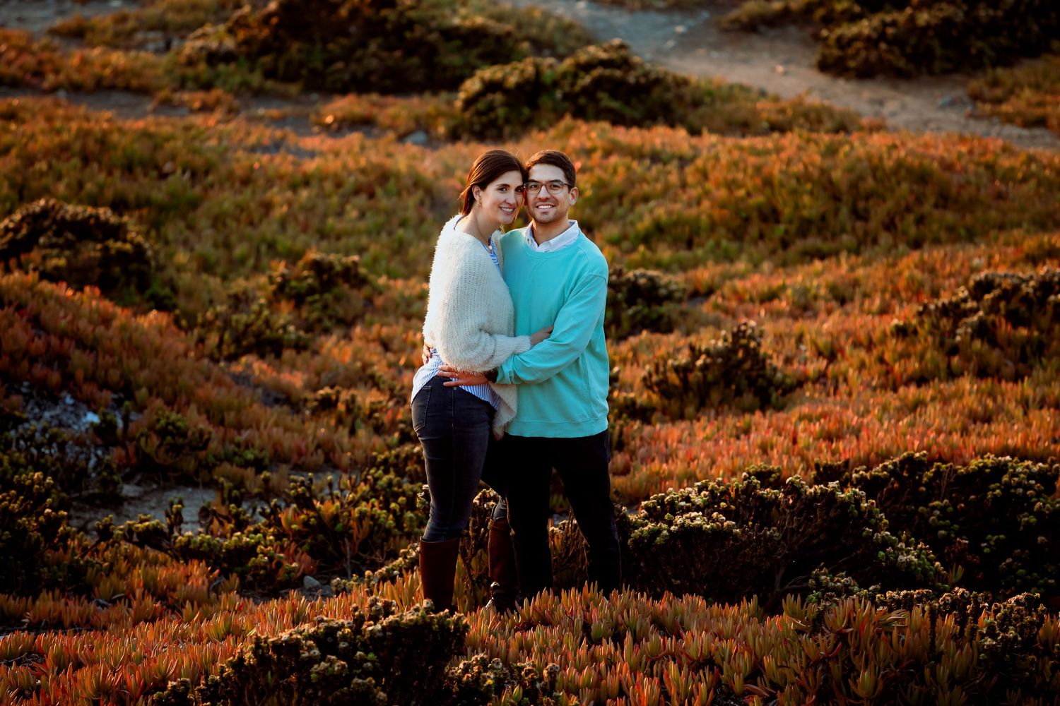 Couple embracing in a vibrant field of autumn-colored coastal vegetation.