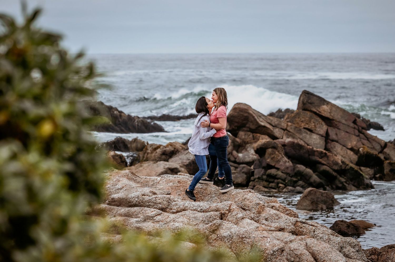 Two women share a romantic embrace on rugged coastal rocks near crashing waves.