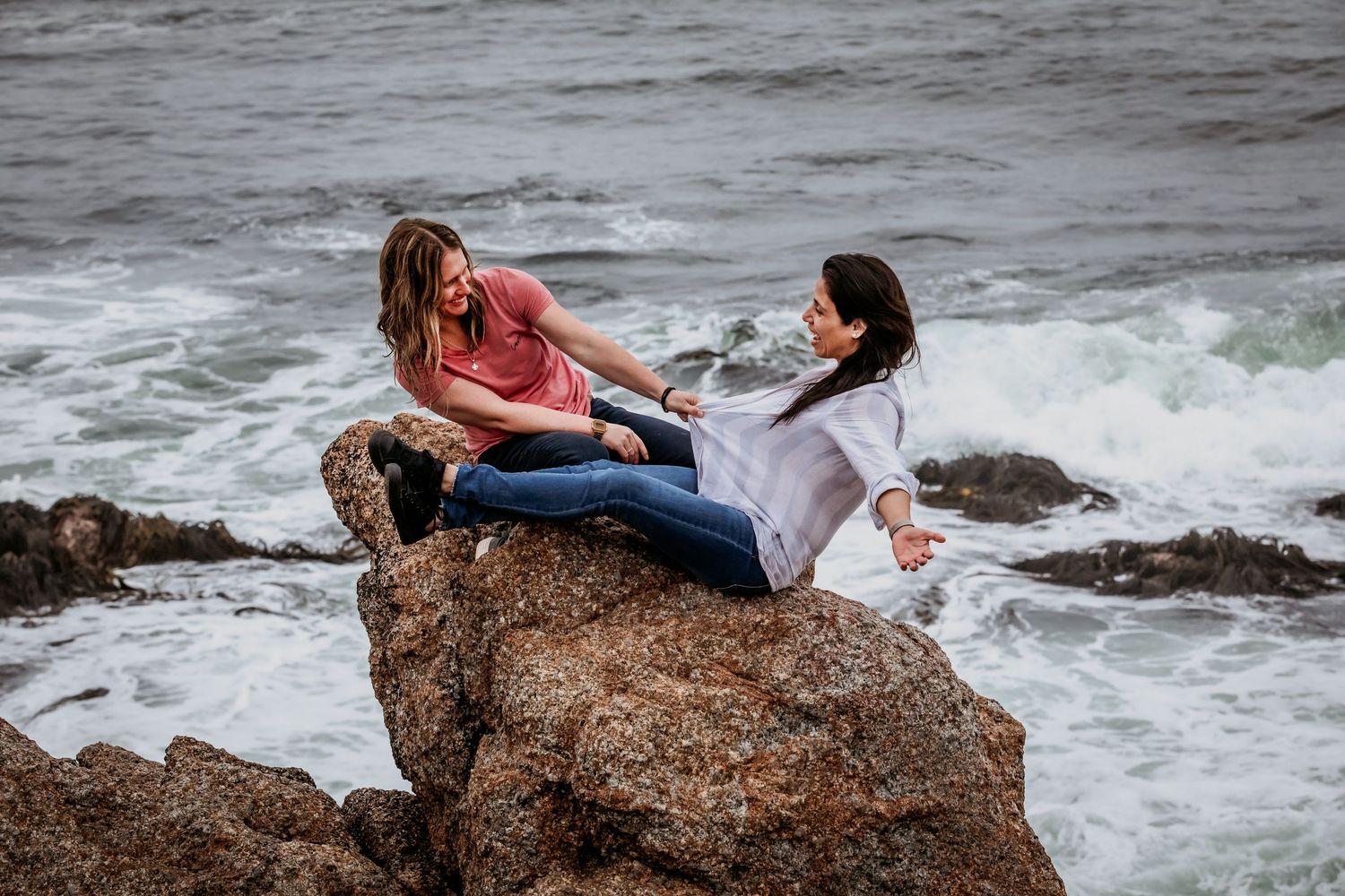 Two women laughing together on rocky shoreline with turbulent ocean waves in background.