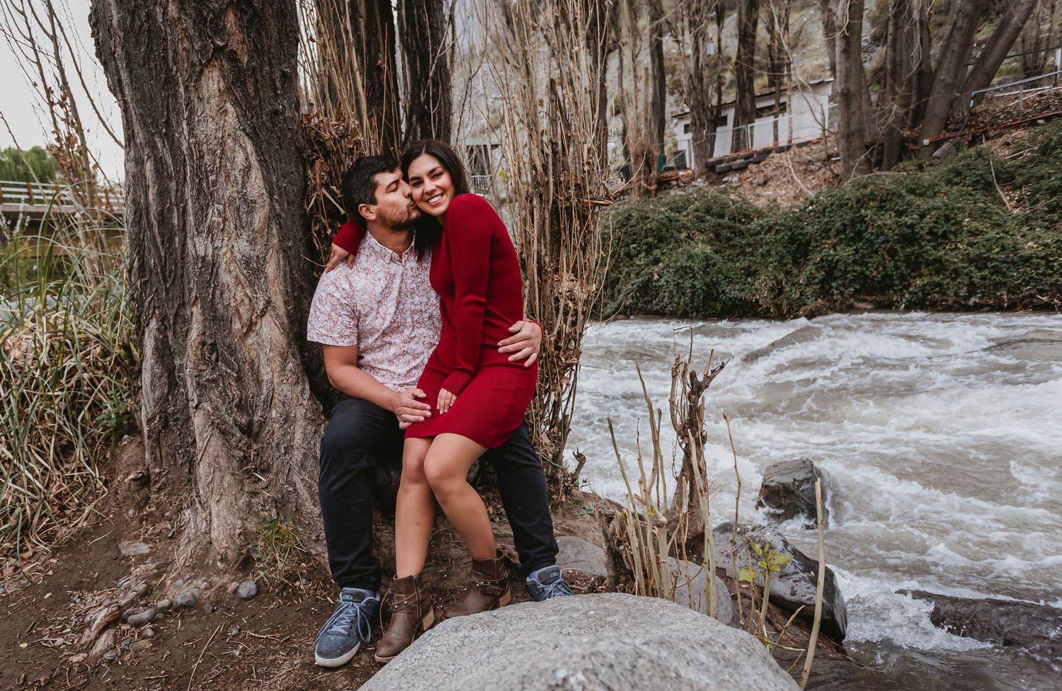 Couple cuddling against tree trunk beside rushing river in early spring outdoor engagement session.