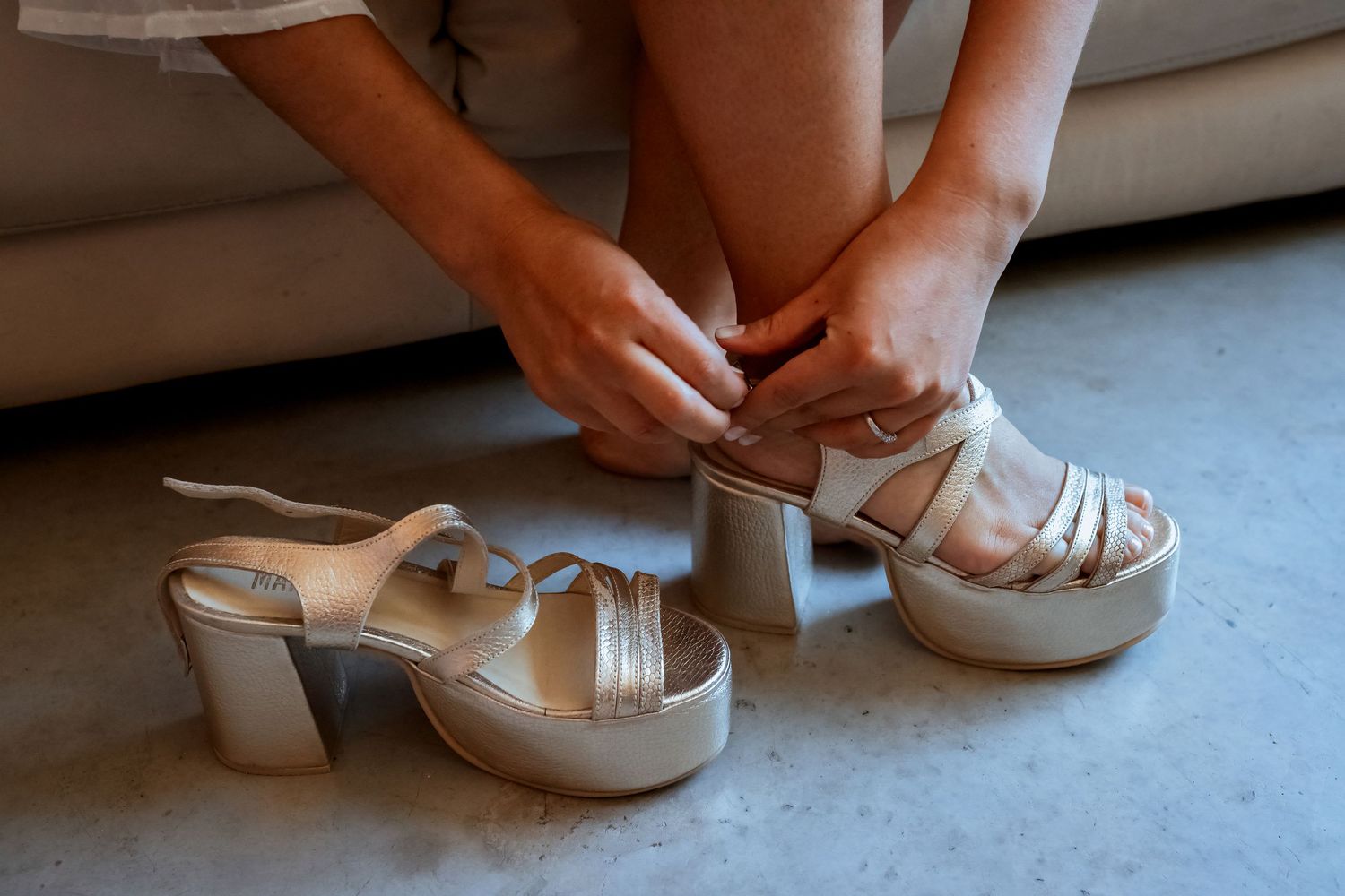 Bride putting on strappy gold platform heels while sitting on a couch during wedding day preparations.