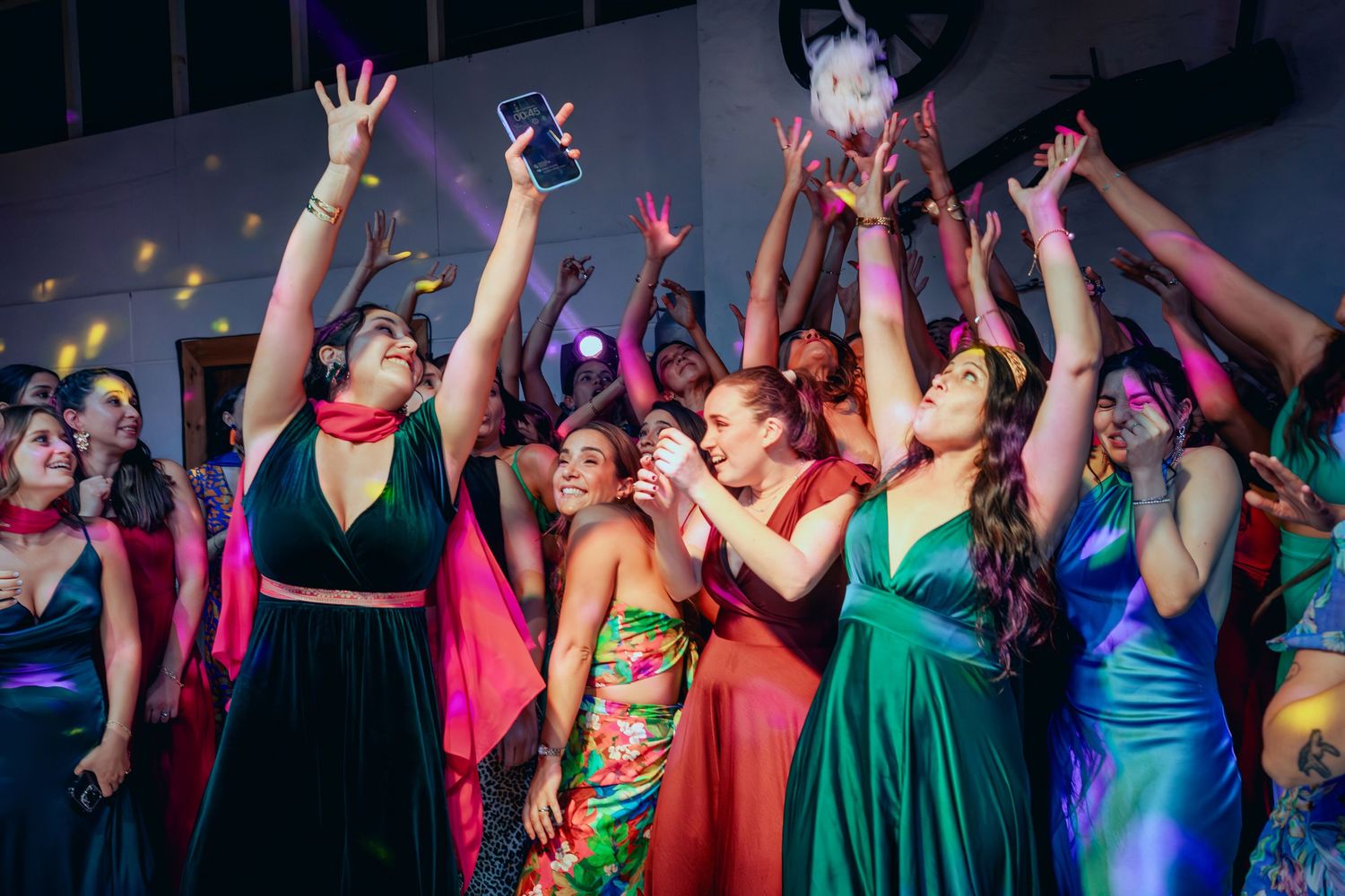 Wedding guests in colorful gowns reach up to catch bridal bouquet toss.