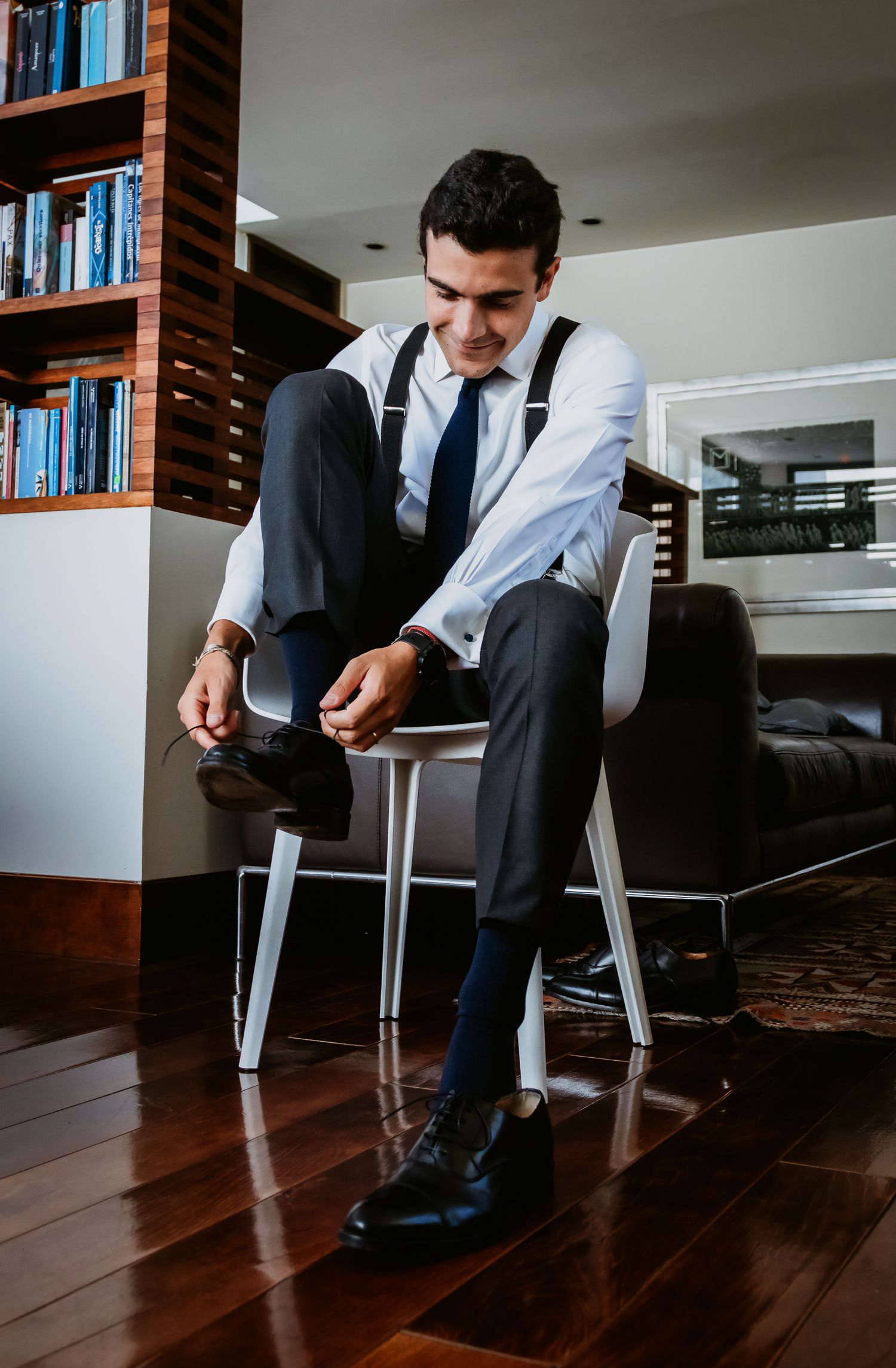 Groom sitting on chair putting on dress shoes wearing suspenders white shirt and navy tie.