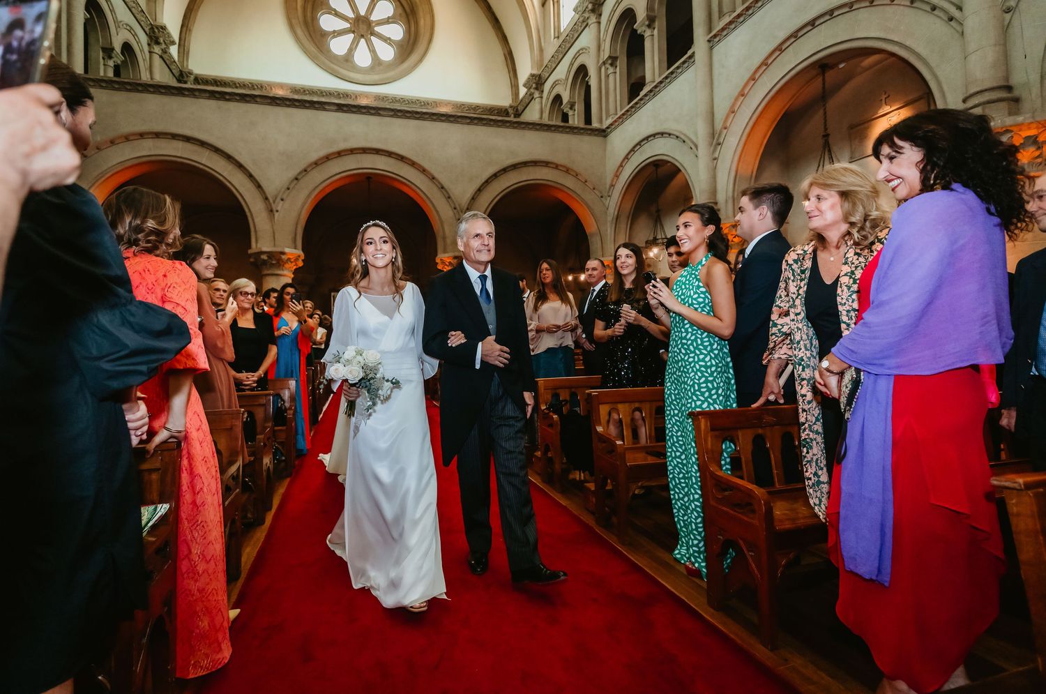 Bride and escort processing down red carpet aisle in grand church with arched stone architecture.